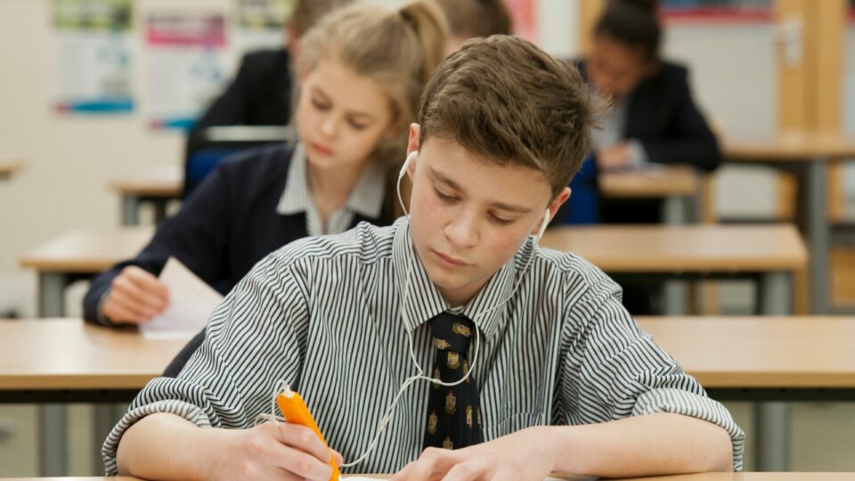 a student sitting at the desk with ExamReader pen in the right hand and headphones with other students in the background